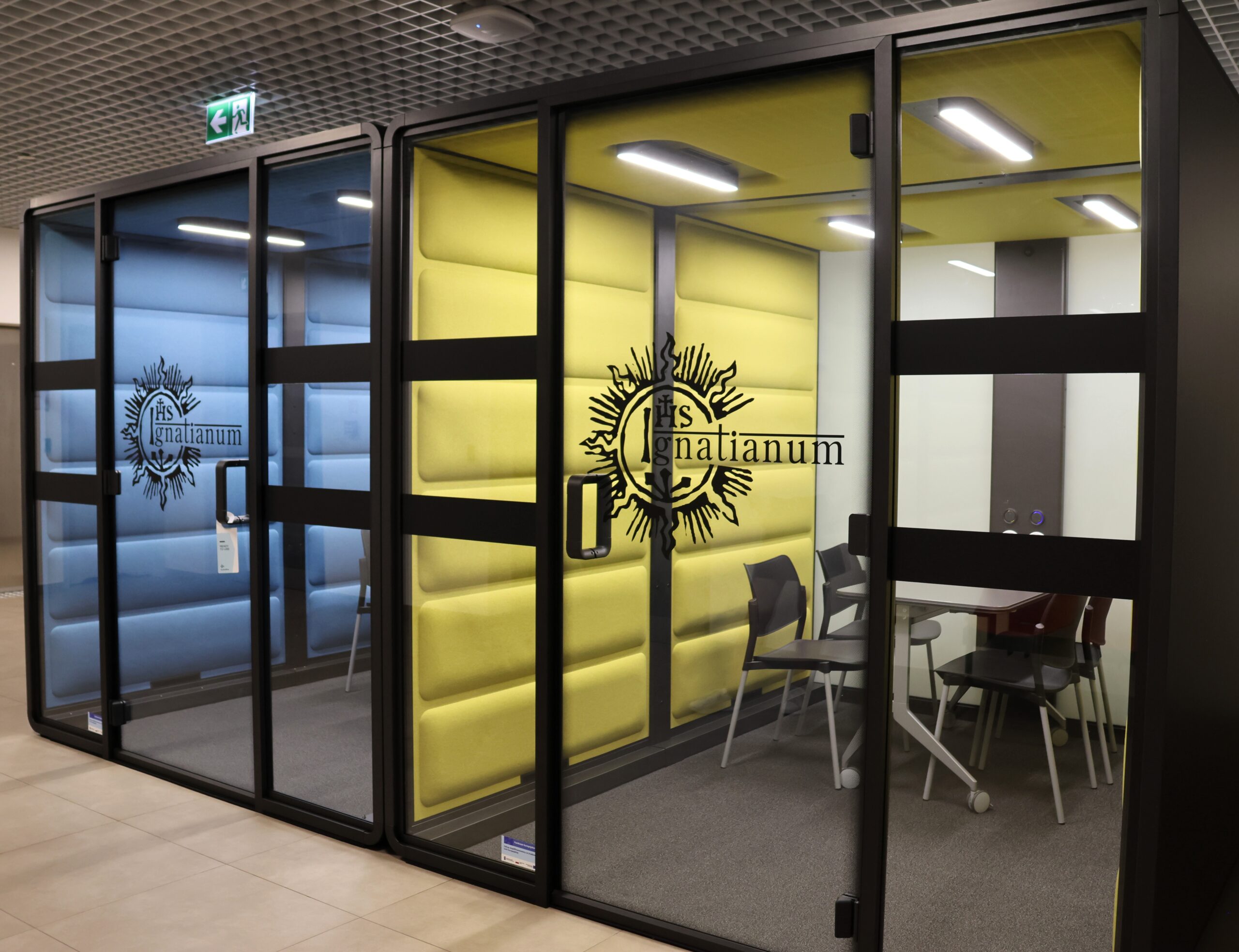 Grafika przedstawia budki wyciszające w kolorze ziebieskim i żółtym z logo Uniwersytetu Ignatianum